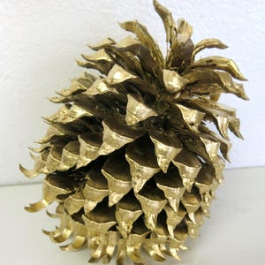 Puede incluir: Una piña grande pintada de oro. La piña es de un color dorado metálico cálido, con cada escama y punta claramente definida. La piña es un artículo decorativo, adecuado para su exhibición.