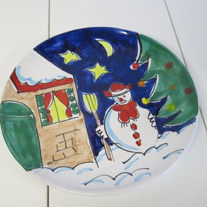 May include: A white ceramic plate with a hand-painted winter scene. The scene features a snowman holding a broom, a house with a red roof, a green tree, and a blue night sky with stars and a crescent moon.