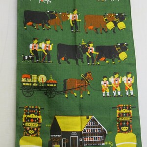 Op de afbeelding: Een groene theedoek met een kleurrijke illustratie van een traditionele Duitse dorpssc&egrave;ne. De sc&egrave;ne bevat koeien, geiten, een paard en wagen, een boerderij en mensen in traditionele kleding. De tekst "G'S'G'T" staat op een bel die bovenaan de afbeelding hangt.