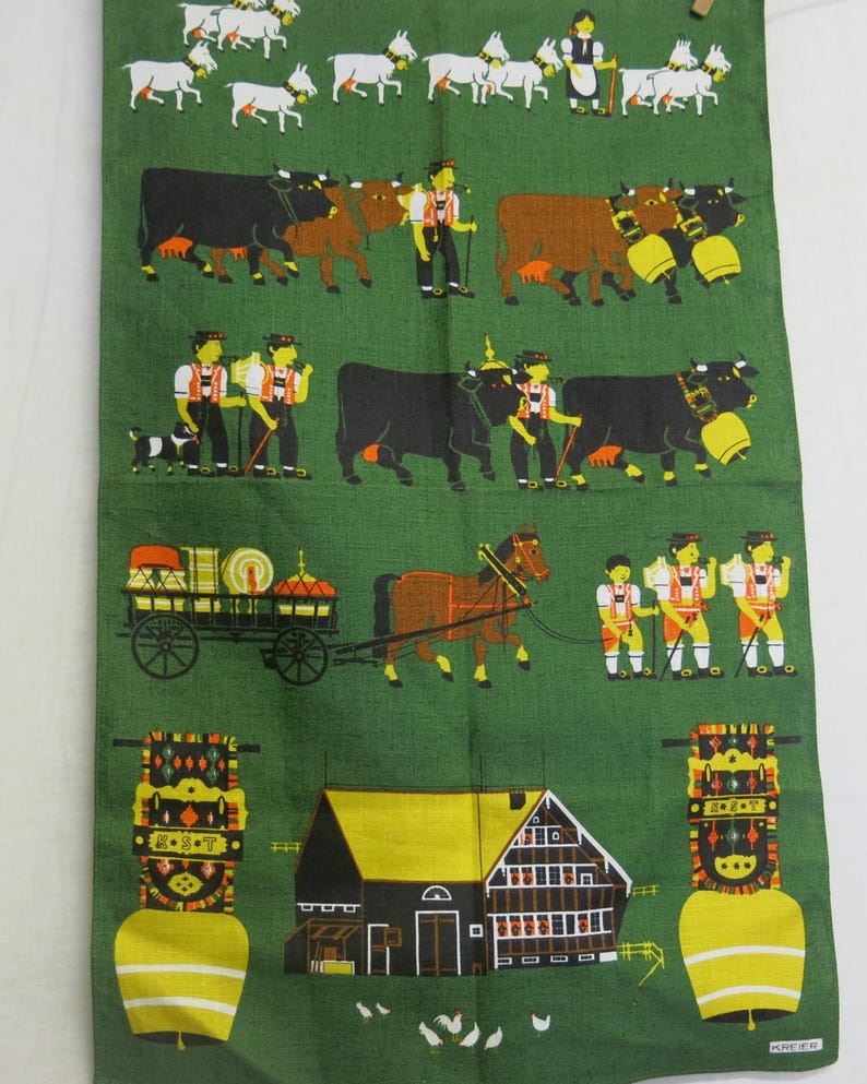 Op de afbeelding: Een groene theedoek met een kleurrijke illustratie van een traditionele Duitse dorpssc&egrave;ne. De sc&egrave;ne bevat koeien, geiten, een paard en wagen, een boerderij en mensen in traditionele kleding. De tekst "G'S'G'T" staat op een bel die bovenaan de afbeelding hangt.