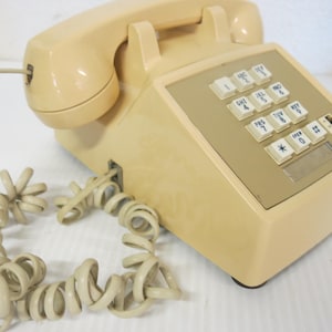 May include: A vintage-style beige landline telephone featuring a push-button keypad with white keys, a matching beige handset, and a coiled cord. The device has a tan-colored faceplate surrounding the numeric buttons and a small rectangular label area.