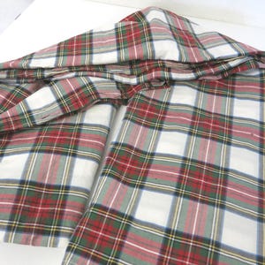 Op de afbeelding: Gevouwen geruite stof in een klassiek patroon van rood, groen, wit en grijs. De stof is in lagen gerangschikt en toont het herhalende vierkante ontwerp. Het materiaal lijkt katoen of een vergelijkbaar geweven textiel te zijn.