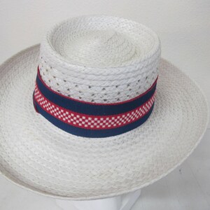 Puede incluir: Un sombrero de paja blanco con una banda a cuadros azul y roja. El sombrero tiene un ala ancha y una textura tejida.