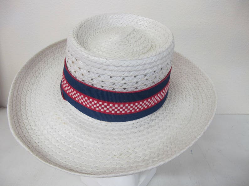 Puede incluir: Un sombrero de paja blanco con una banda a cuadros azul y roja. El sombrero tiene un ala ancha y una textura tejida.