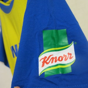 May include: A blue t-shirt with a yellow and blue design. The shirt has the Knorr brand logo on the sleeve.