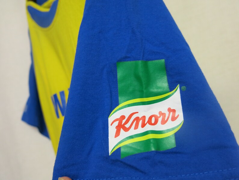 May include: A blue t-shirt with a yellow and blue design. The shirt has the Knorr brand logo on the sleeve.