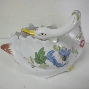 Puede incluir: Jardinera de cerámica blanca con forma de cisne, con pico amarillo y detalles florales. La jardinera presenta un cuello curvo y un borde con volantes, con diseños florales azules, verdes y rosas pintados a mano. La cola del cisne es marrón.