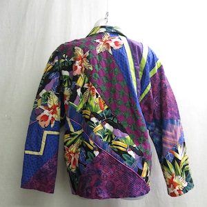 May include: A colorful quilted jacket with a tropical print featuring toucans, flowers, and leaves. The jacket is made of various fabrics in shades of purple, blue, green, and yellow.