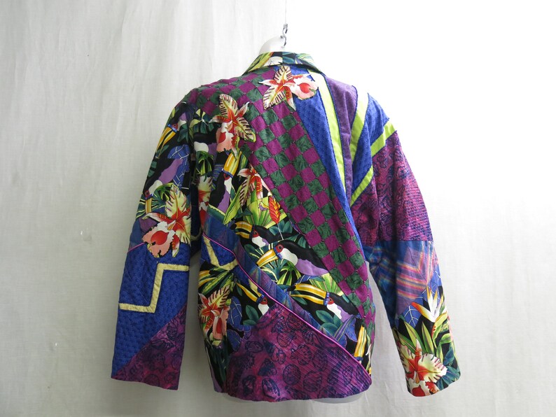 May include: A colorful quilted jacket with a tropical print featuring toucans, flowers, and leaves. The jacket is made of various fabrics in shades of purple, blue, green, and yellow.