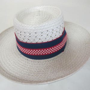 Puede incluir: Un sombrero de paja blanco con una banda a cuadros azules y rojos. El sombrero tiene un ala ancha y est&aacute; hecho de un material tejido.