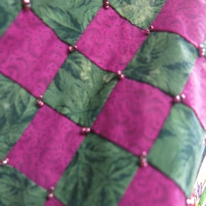 May include: A close-up of a pink and green patchwork quilt with a checkered pattern. The quilt is decorated with small, round beads.