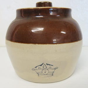 May include: A two-toned stoneware crock with a dark brown glazed upper section and a light beige lower section. The vessel features a lid with a central knob and a blue stamped crown graphic containing the number two on the front exterior.