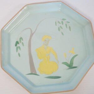 May include: An octagonal ceramic plate with a light blue background and a tan rim. The plate features a stylized illustration of a person in yellow clothing under a tree. The tree has gray bark and green leaves.