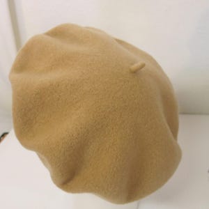 May include: A tan beret hat made of a soft, textured material. The beret is round with a small, cylindrical detail on top. The hat is displayed on a white mannequin head against a white background. This classic hat is a stylish accessory.
