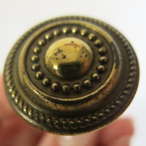 May include: A gold-toned metal knob with a raised, circular design. The knob has a beaded border and a rope-like edge.