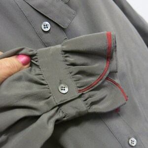 Op de afbeelding: Grijze zijden blouse met een gerimpelde manchet en een rode bies. De manchet heeft een knoopsluiting.