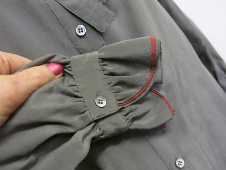 Op de afbeelding: Grijze zijden blouse met een gerimpelde manchet en een rode bies. De manchet heeft een knoopsluiting.