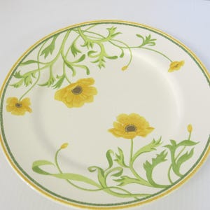 May include: A round, off-white plate with a yellow and green floral design. The plate features yellow flowers with brown centers and green leaves and stems. A yellow and green border frames the edge of the plate.