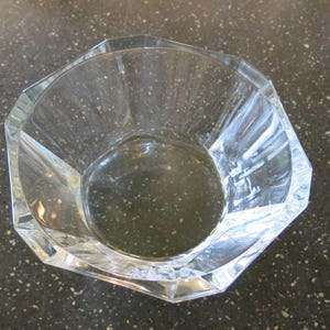 Puede incluir: Un cuenco de vidrio transparente y facetado con forma octogonal. El cuenco tiene una abertura ancha y una base plana. El vidrio es transparente, lo que permite el paso de la luz. El cuenco se utiliza probablemente para servir o exhibir.