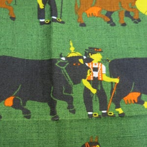 Op de afbeelding: Een groene stof met een herhalend patroon van zwarte en bruine koeien, een man in traditionele kleding en een paard. De man houdt een stok vast en het paard draagt een rood hoofdstel.