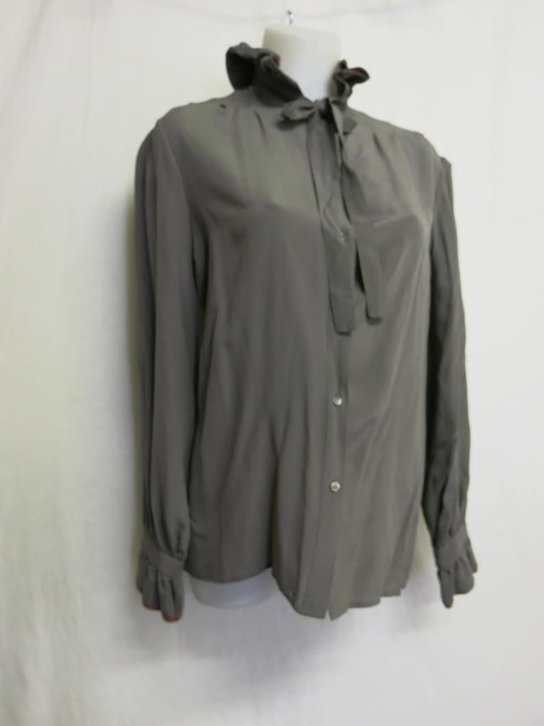 Op de afbeelding: Een grijs, lang mouwig blouse met een gerimpelde kraag en een strik. De blouse heeft een knoopsluiting aan de voorkant en een aansluitende pasvorm.