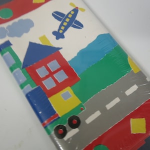 May include: A roll of colorful wallpaper with a repeating pattern of a house, a plane, and a road. The house is red, blue, green, and yellow. The plane is blue and yellow. The road is gray with white lines. The background is white.