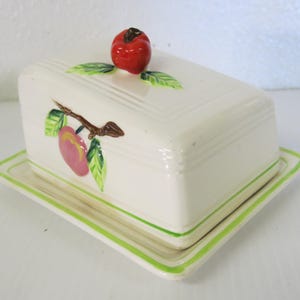 Può includere: Una burriera rettangolare in ceramica bianco sporco con coperchio, decorata con una mela rossa e foglie verdi sulla parte superiore. Il piatto presenta un disegno di pesca e foglie e un bordo verde. Un affascinante articolo per la cucina.