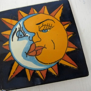 May include: A pair of ceramic tiles with a blue background and a painted image of the sun and moon kissing. The sun is orange and the moon is white with blue highlights. The sun and moon have eyes and lips.