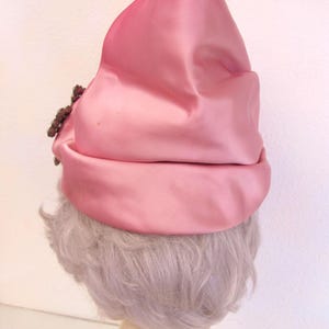 Op de afbeelding: Een roze satijnen hoed met een afgeronde kroon en een omgeslagen rand. Een cluster van donkerbruine kralen siert de zijkant. De hoed heeft een zachte, gedempte roze kleur en de stof heeft een subtiele glans.
