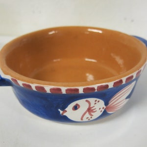 May include: A small blue ceramic bowl with a red and white rim. The bowl has a painted design of a white fish with pink fins on the side.