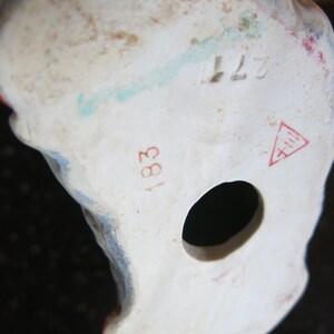May include: Close-up of the base of a white porcelain figurine. The figurine has a hole and several markings, including the numbers "183" and "277", and a red triangle symbol. The figurine has blue and red paint details.