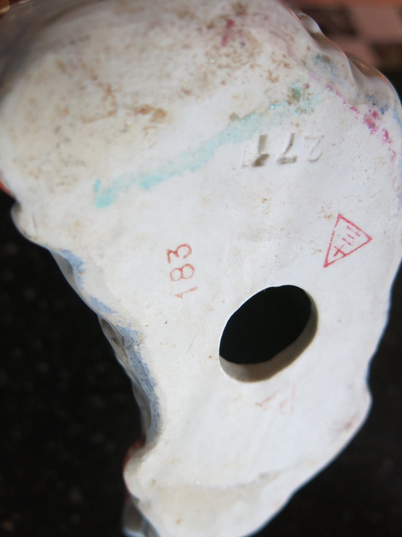May include: Close-up of the base of a white porcelain figurine. The figurine has a hole and several markings, including the numbers "183" and "277", and a red triangle symbol. The figurine has blue and red paint details.