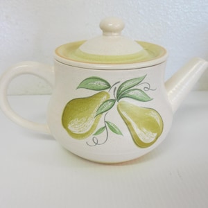 Puede incluir: Tetera de cerámica color crema con tapa y asa. La tetera está decorada con peras y hojas verdes. La tapa tiene una banda verde claro. La tetera tiene un pico para verter.