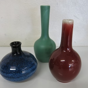 May include: Three small ceramic vases in different colors and styles. The vase on the left is blue with a speckled glaze. The vase in the center is green and has a simple, rounded shape. The vase on the right is red with a glossy finish.
