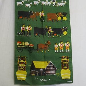 Op de afbeelding: Een groene theedoek met een kleurrijke illustratie van een traditionele Beierse sc&egrave;ne. De sc&egrave;ne bevat koeien, geiten, een paard en wagen, een boerderij en mensen in traditionele kleding. De theedoek heeft een groene achtergrond met gele, oranje, rode en zwarte accenten.
