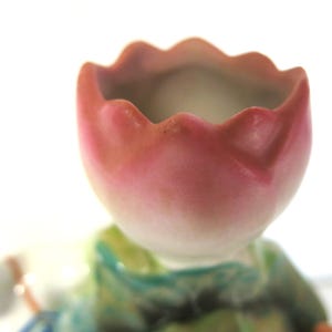 May include: A close-up of a ceramic figurine featuring a pink and white tulip-shaped head. The figurine has a green and blue outfit. The background is white.