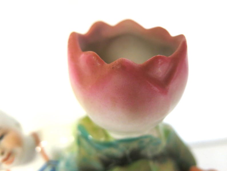 May include: A close-up of a ceramic figurine featuring a pink and white tulip-shaped head. The figurine has a green and blue outfit. The background is white.