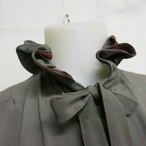 Op de afbeelding: Een grijs blouse met een gerimpelde kraag en een strik. De kraag heeft een rode rand.