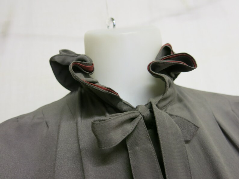 Op de afbeelding: Een grijs blouse met een gerimpelde kraag en een strik. De kraag heeft een rode rand.