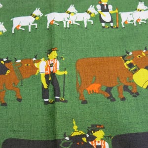 Op de afbeelding: Een groene stof met een kleurrijke illustratie van mensen en dieren. De illustratie toont een groep mensen in traditionele kleding, koeien en geiten. De stof heeft een textuur.
