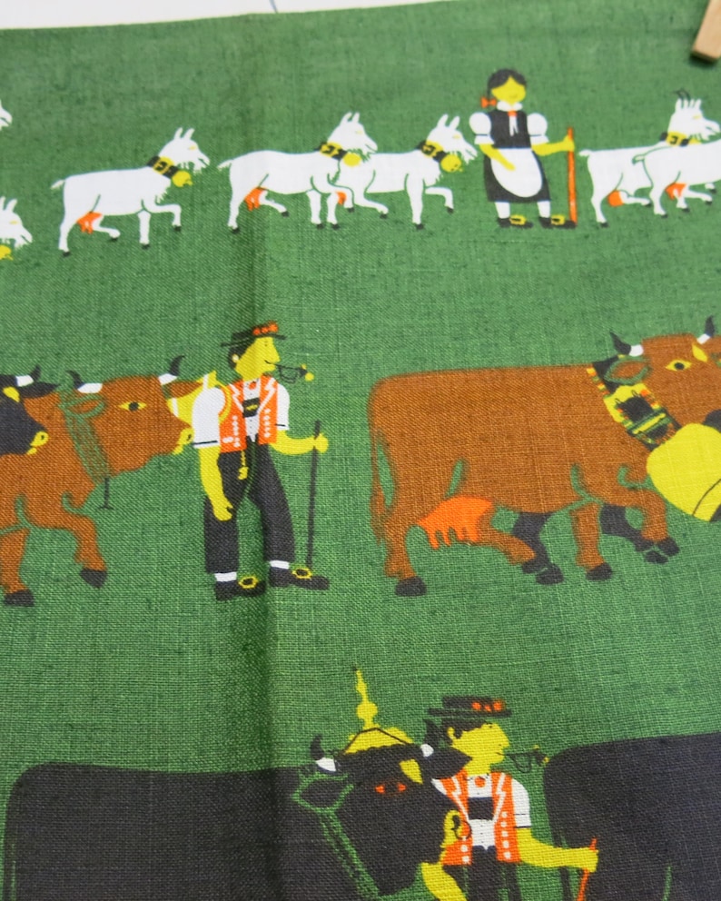 Op de afbeelding: Een groene stof met een kleurrijke illustratie van mensen en dieren. De illustratie toont een groep mensen in traditionele kleding, koeien en geiten. De stof heeft een textuur.