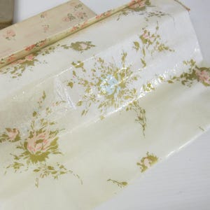 May include: A roll of off-white paper featuring a repeating floral pattern of light pink roses and olive green foliage. The paper has a glossy finish and is partially unrolled from a rectangular box with matching floral print and the text "Perfume".