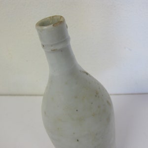Könnte beinhalten: Eine weiße Keramikflasche mit einem runden Körper und einem schmalen Hals. Die Flasche hat eine leicht strukturierte Oberfläche und scheint vintage zu sein.