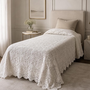 May include: A cream-coloured bedspread featuring an intricate, repeating floral crochet pattern with a scalloped edge. The textile covers a single bed, draping over the sides and foot, showcasing a detailed, openwork knit design throughout the fabric.