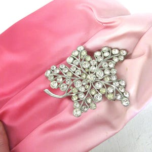 Op de afbeelding: Een vintage roze satijnen clutch met een zilverkleurige, met strass bezette bladbroche. De clutch heeft een tweekleurig roze ontwerp, met een donkerdere tint aan de ene kant en een lichtere tint aan de andere kant.