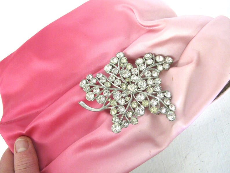 Op de afbeelding: Een vintage roze satijnen clutch met een zilverkleurige, met strass bezette bladbroche. De clutch heeft een tweekleurig roze ontwerp, met een donkerdere tint aan de ene kant en een lichtere tint aan de andere kant.