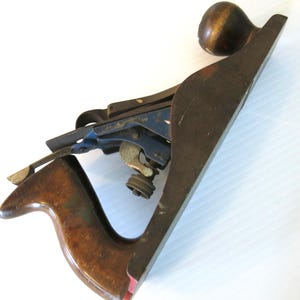 May include: A vintage hand plane with a wooden body and handle, featuring a dark finish. The plane has a metal blade and adjustment mechanism, with a brass-colored knob. The tool is designed for woodworking and shaping materials.