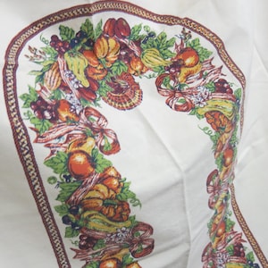 Puede incluir: Tela blanca con un borde decorativo con un diseño de temática de cosecha. El borde incluye ilustraciones coloridas de calabazas, calabacines, uvas y lazos. El diseño está enmarcado por un borde marrón estampado.