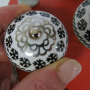May include: Close-up of a white ceramic knob with a black and silver floral design. The knob has a silver metal screw in the center.