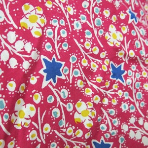 May include: Pink fabric with a white floral pattern. The flowers have yellow centers and are surrounded by blue stars. The fabric is textured and has a slightly wrinkled appearance.